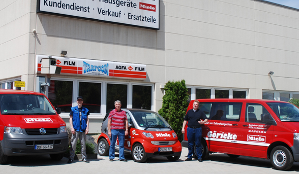 Drei rote Firmenfahrzeuge mit drei Personen vor einem Firmengebäude