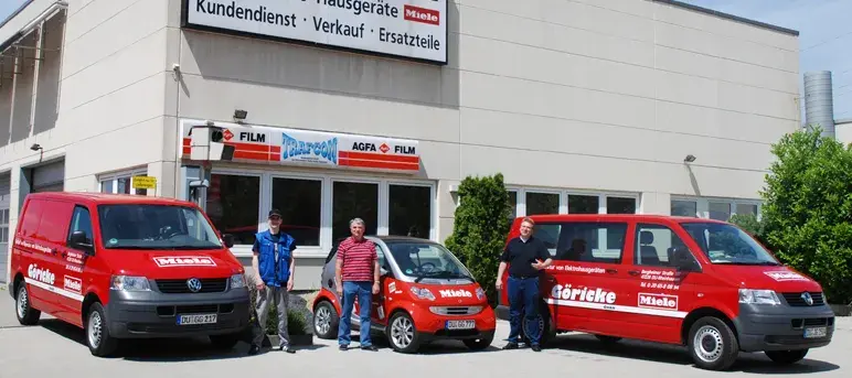 Drei rote Firmenfahrzeuge mit drei Personen vor einem Firmengebäude